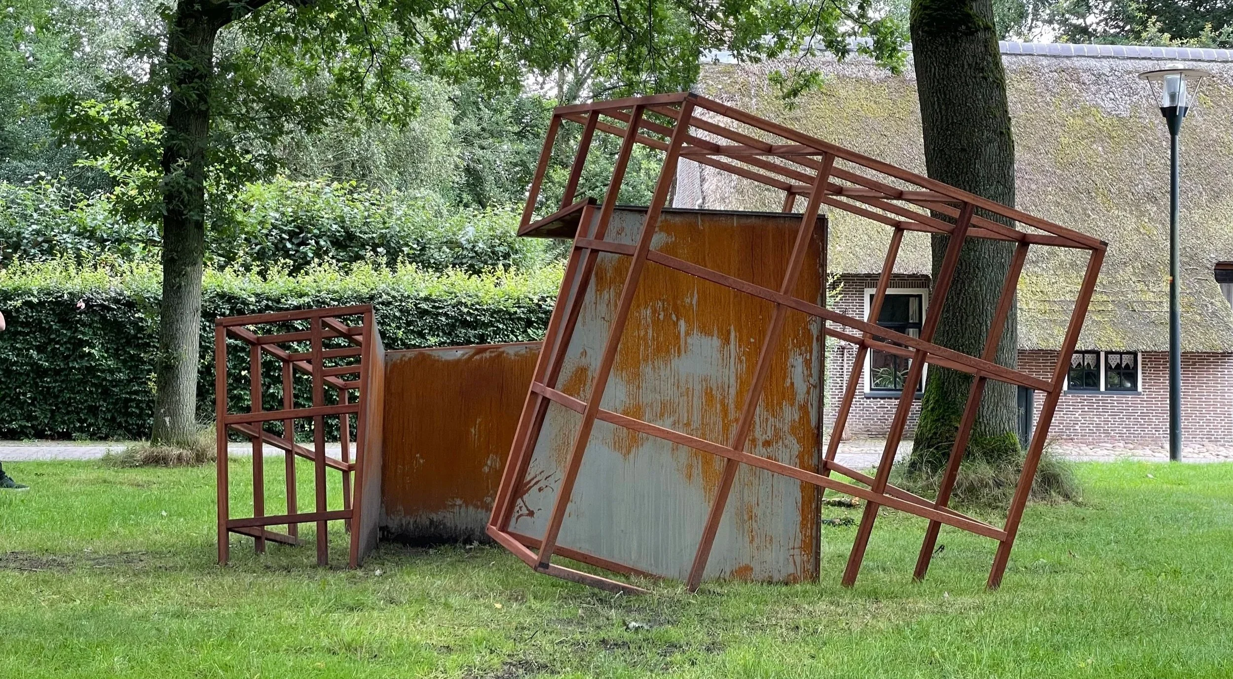'Betekenisloos' kunstwerk bestaande uit een houten frame met metalen platen, neergezet op een grasveld onder bomen. Het kunstwerk verbeeldt drie kubussen die in verval zijn, en verwijst naar de tijdelijkheid en fragiliteit van vrede en vrijheid