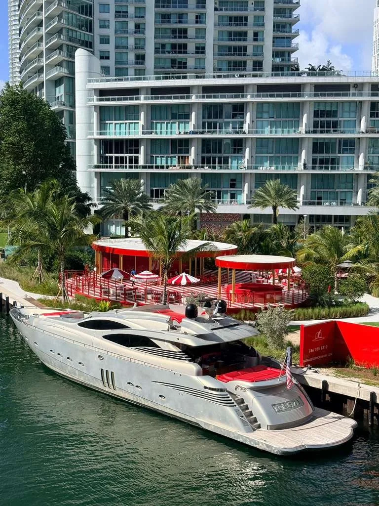 Luxury yacht docked along a waterfront with a modern high-rise building and palm trees in the background. Red and white shaded seating area on the outdoor deck.