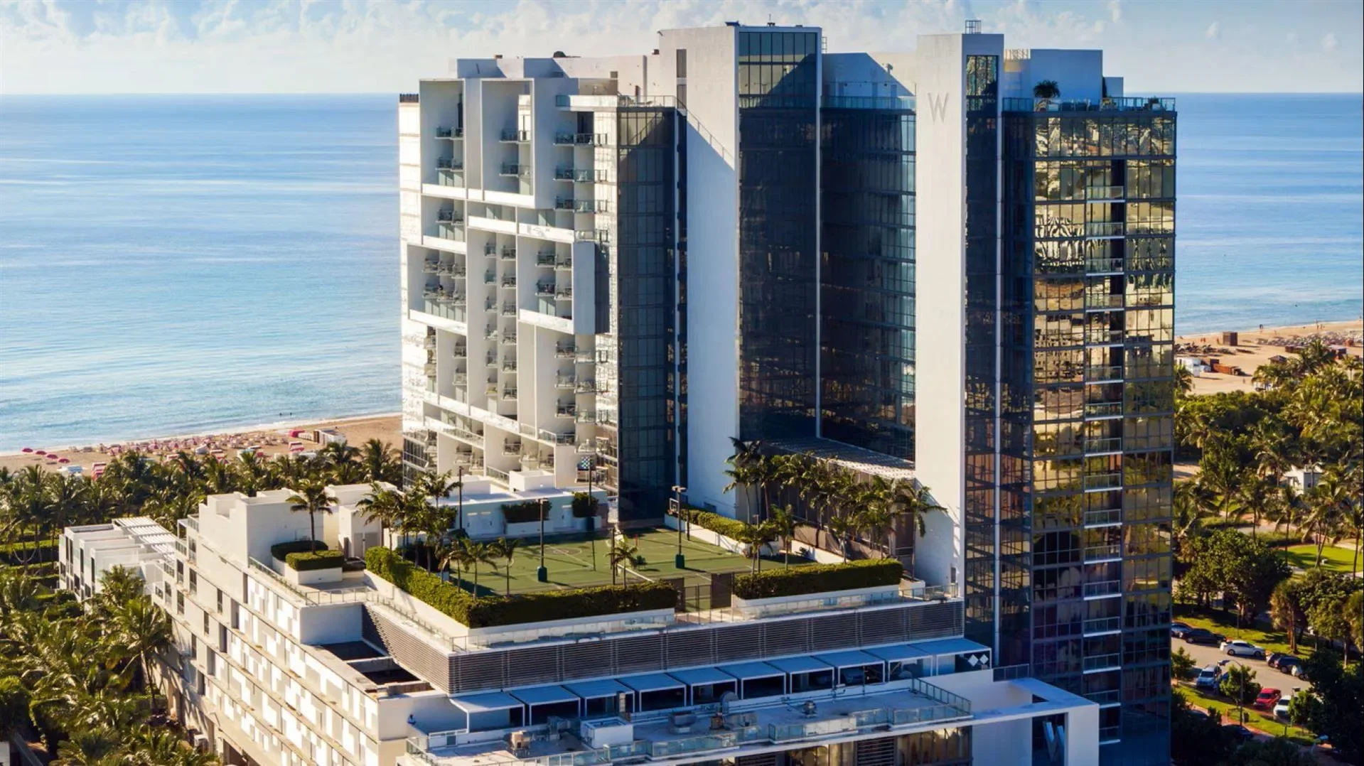 A modern high-rise condominium building with a rooftop tennis court, located along a beach with sandy shore and ocean in the background, surrounded by palm trees.