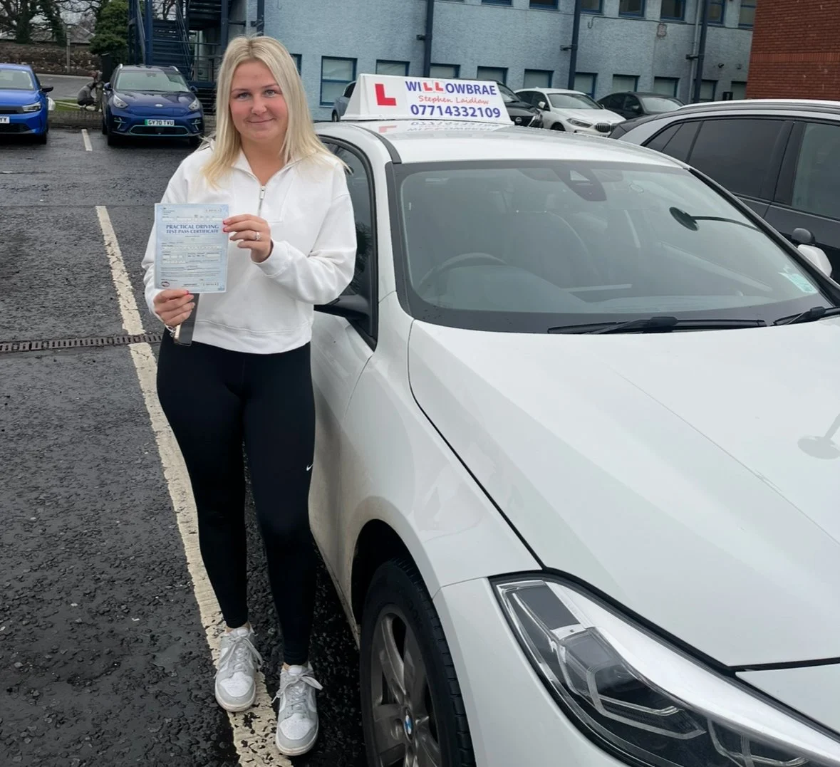 A young woman holding a practical driving test pass certificate standing next to a white driving school car with an instructor's sign on the roof in a parking lot.