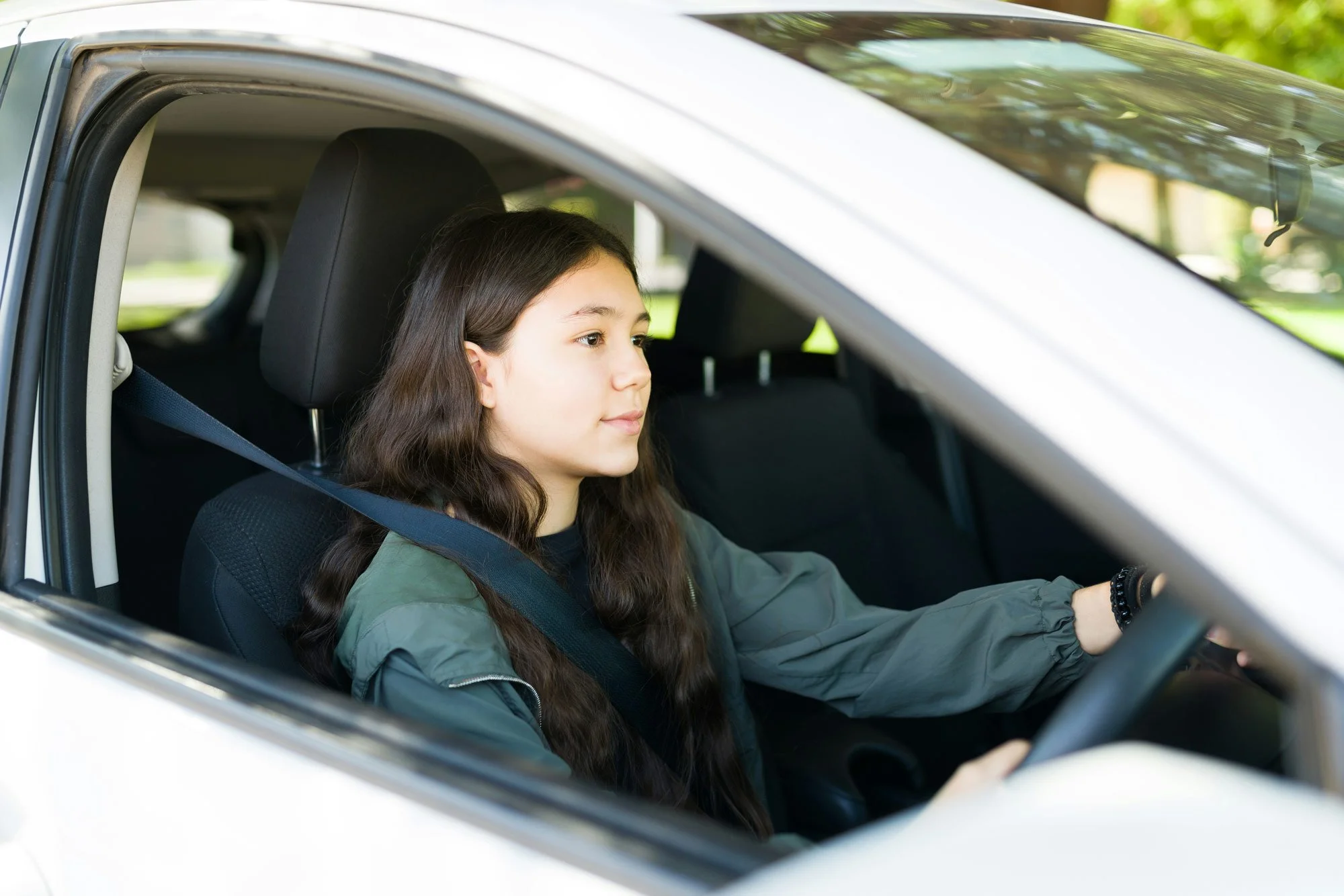 A young girl with long brown hair driving a silver car, wearing a green jacket and a seatbelt.