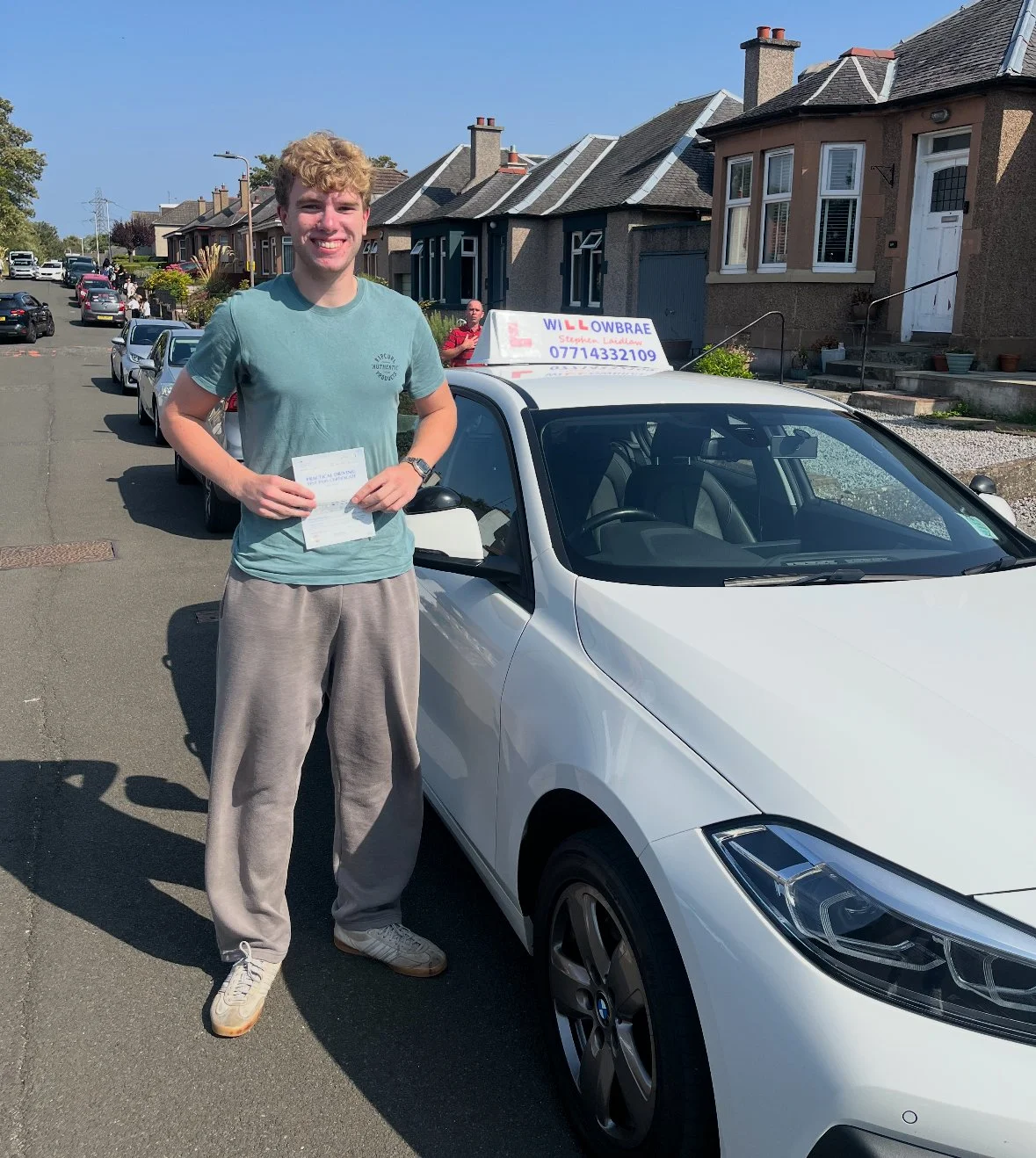 A young man standing next to a white driving school car on a residential street, holding a booklet, smiling, with a sign on the car's roof displaying the name 'Willowbrae' and contact information.