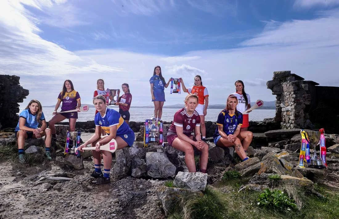 Team of women in the Ireland Women's Rubgy team kit, some sitting and some standing, holding trophies and sporting award cups on a rocky coast before a dramatic backdrop of the blue sky and the sea.
