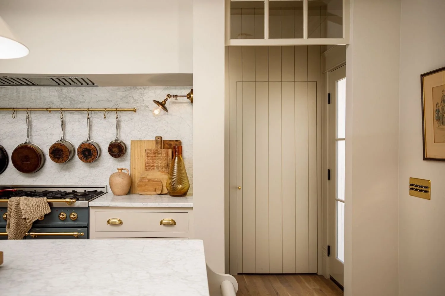 A cozy kitchen corner featuring hanging copper pots, a beige vase, kitchen cutting boards, and a vintage-style stove with gold accents, with a white marble backsplash and a doorway leading outside.