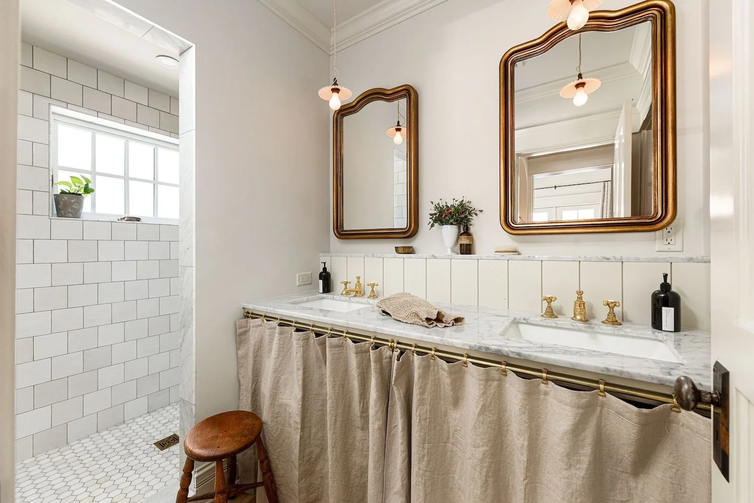 Bathroom with double vanity with marble countertop, gold fixtures, and mirrors, and a separate shower area with white subway tiles and a small window.