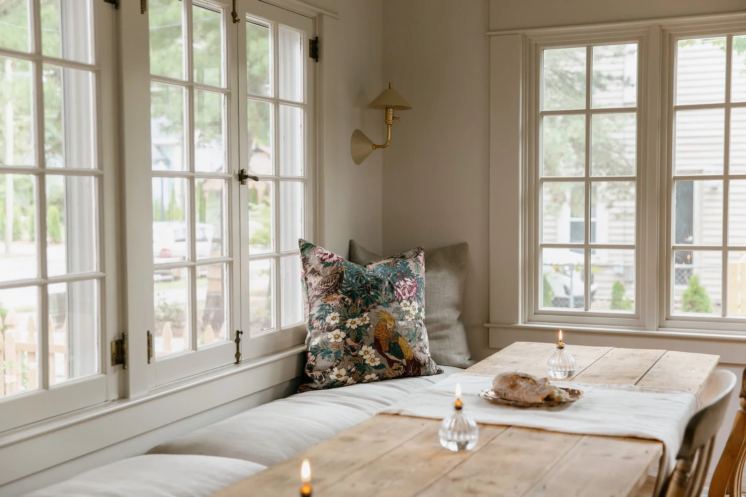 A cozy nook by large windows with decorative pillows on a built-in window seat, a rustic wooden table with a cloth, candles, and a small lamp, set in a bright room with white walls and siding outside.