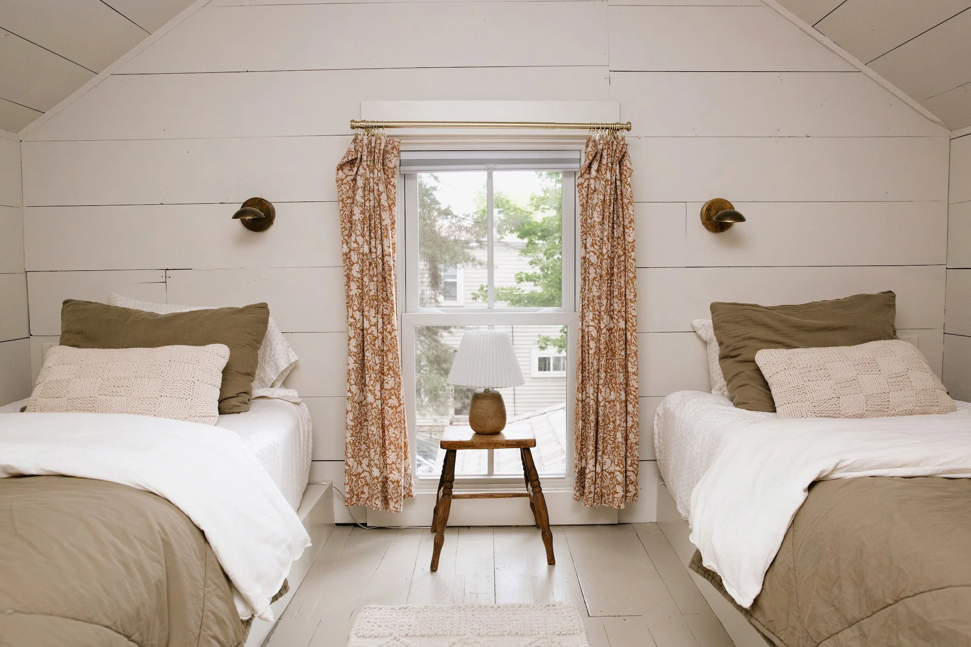 Room with two beds separated by a window and a small wooden table with a lamp, beige bedding and pillows, white walls, and vintage wall sconces.