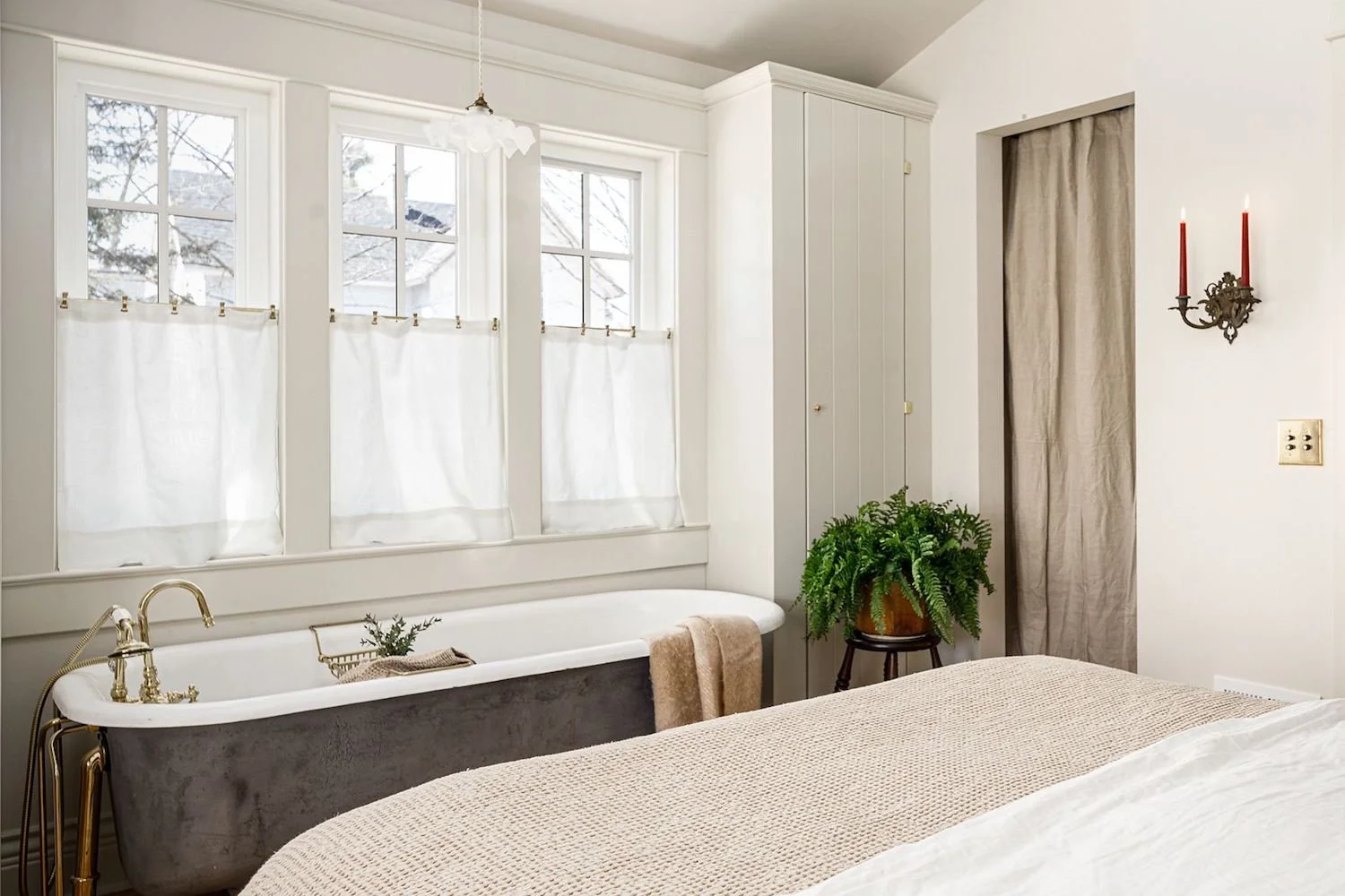 A cozy bedroom with a vintage bathtub by the window, beige bedspread, potted plant on a stand, and a wall sconce with red candles.