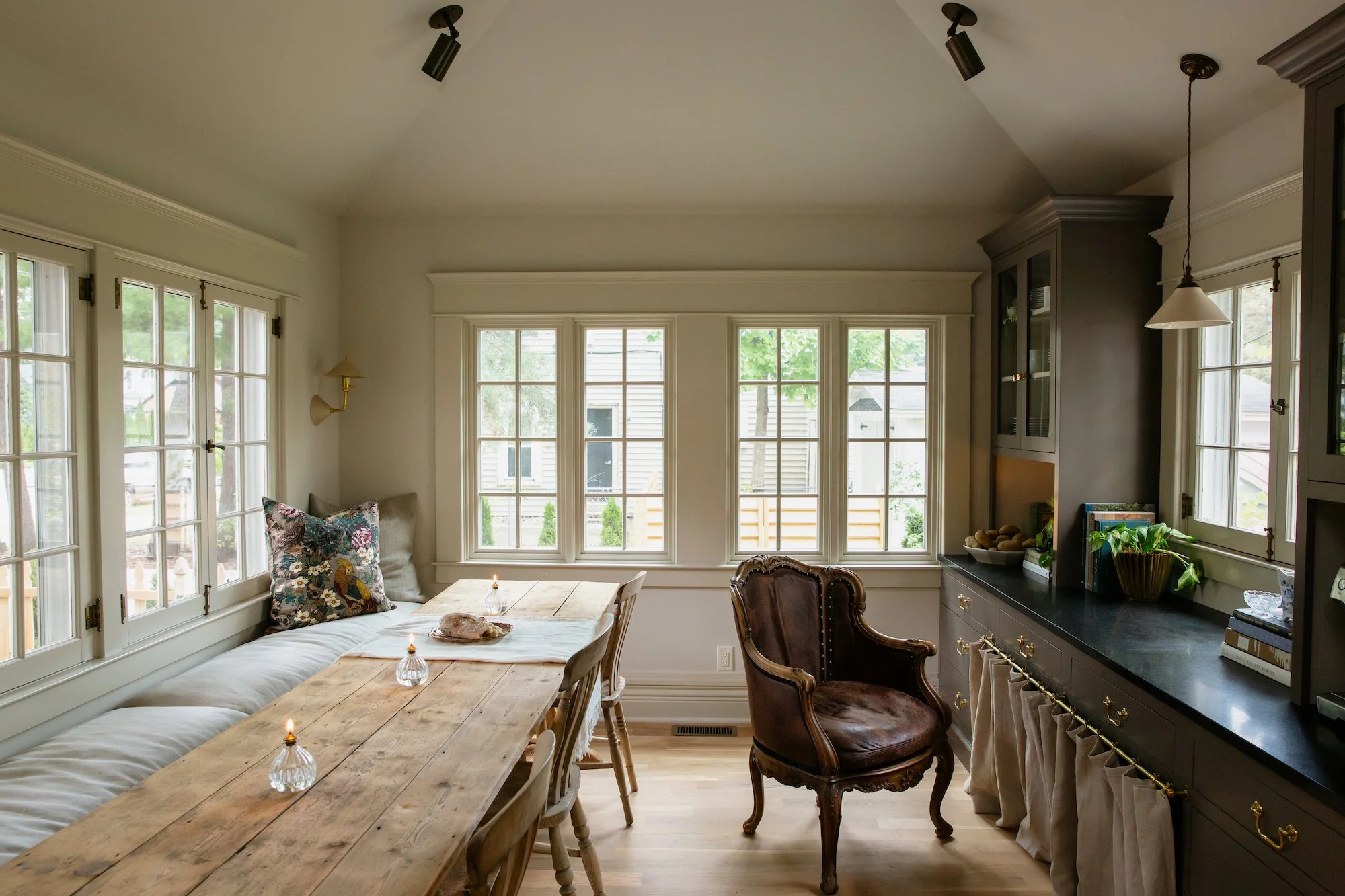 Bright dining room with a large wooden table, vintage armchair, built-in window seat with cushions, and multiple large windows bringing in natural light.