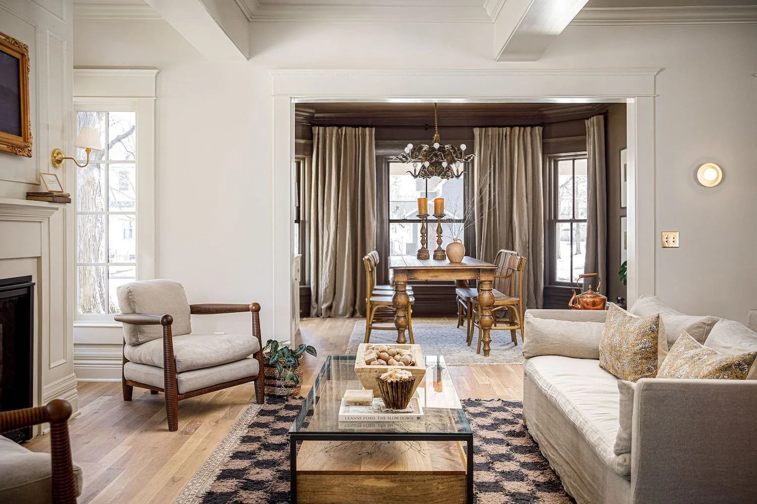 A cozy living room with beige sofa and armchair, glass coffee table, wooden flooring, and a dining area with a wooden table and chairs, large windows with curtains, and a chandelier.