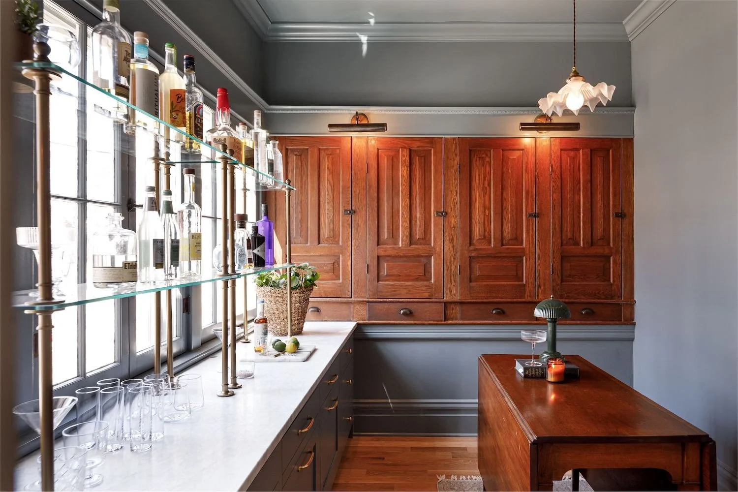 A cozy kitchen with wooden cabinets, a marble countertop, and a shelf with various bottles of liquor. There are glasses on the counter and a potted plant. A window allows natural light to fill the space. A hanging light fixture and a small table with