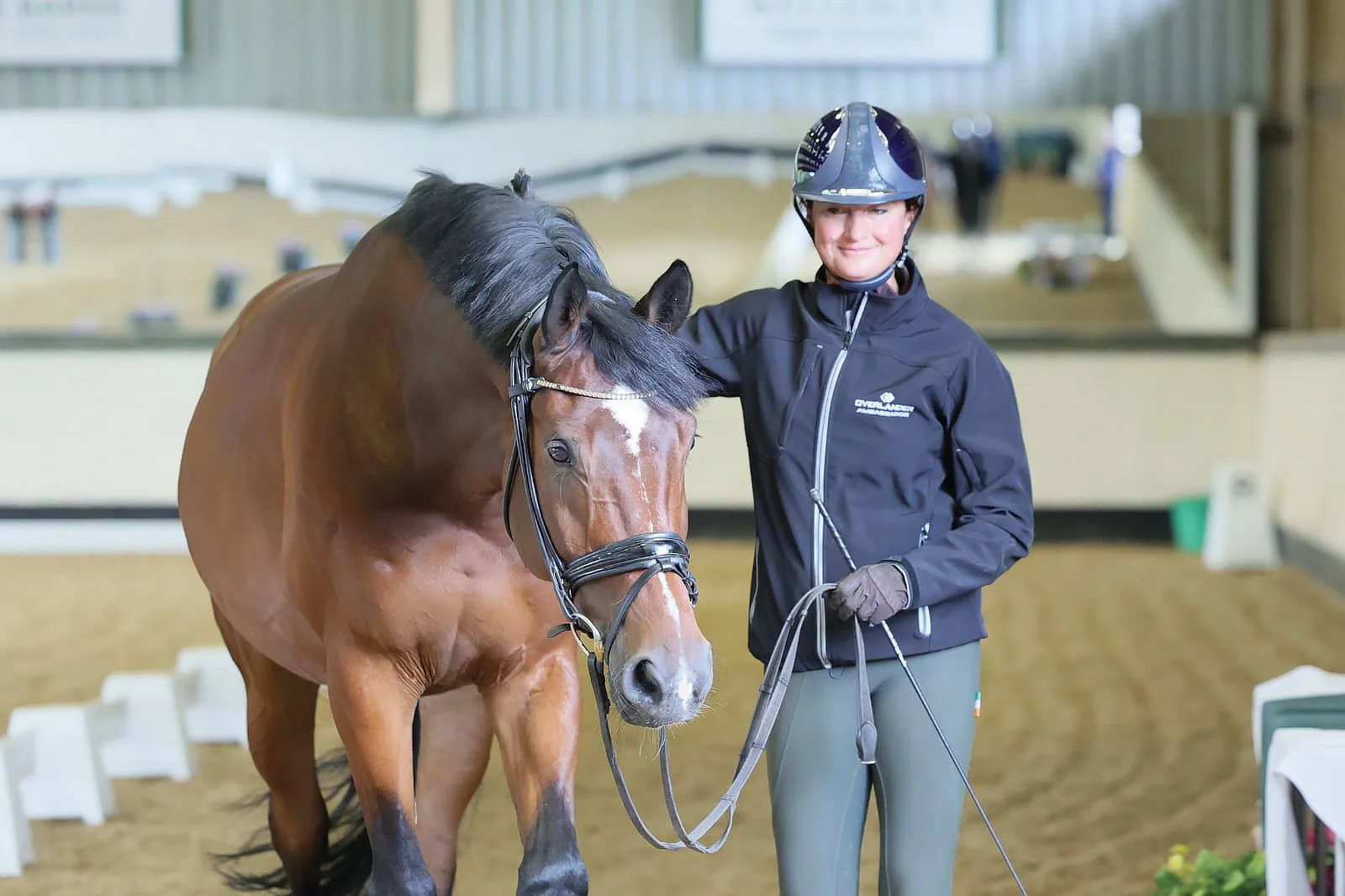 Sorrell Klatzko wearing a helmet and a green Ireland shirt with a braid in her hair is adjusting a brown horse with a bridle.