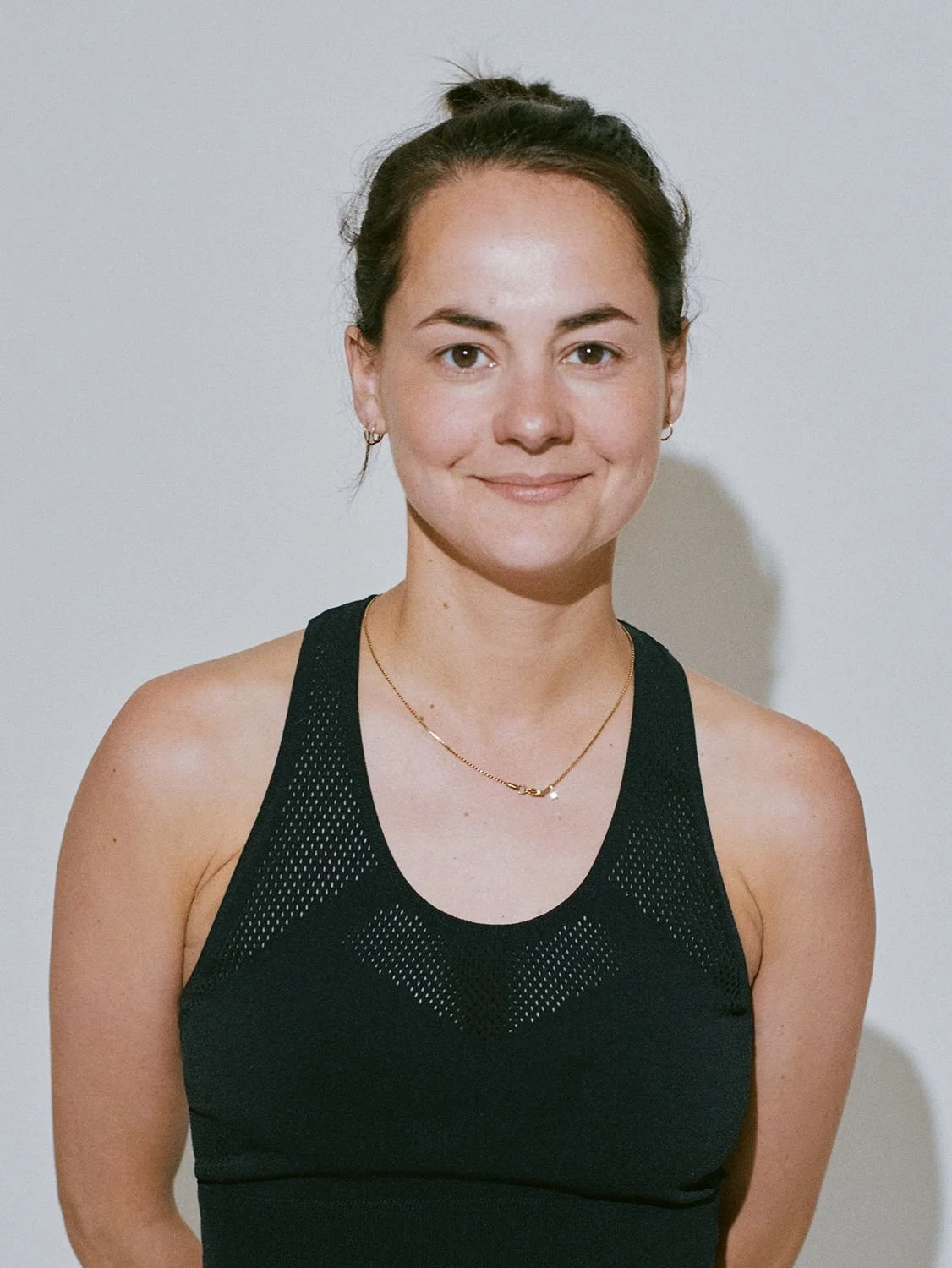 A young woman with dark hair in a bun, wearing a black athletic top with mesh details, gold necklace, and small hoop earrings, standing against a plain light-colored wall.