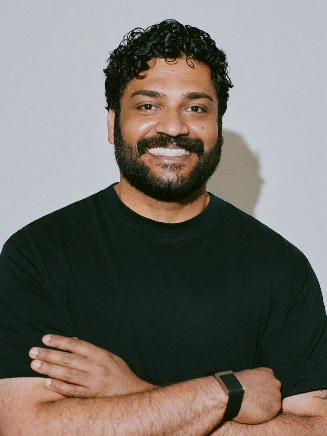Portrait of a smiling man with curly black hair, a beard, wearing a black T-shirt, arms crossed, standing against a plain light-colored background.
