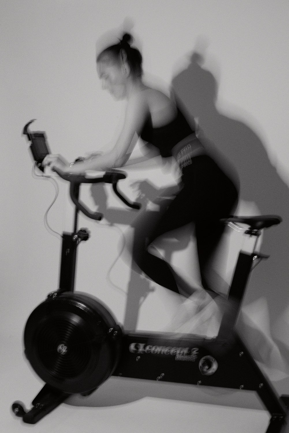 Black and white photo of a woman riding an exercise bike against a plain wall.
