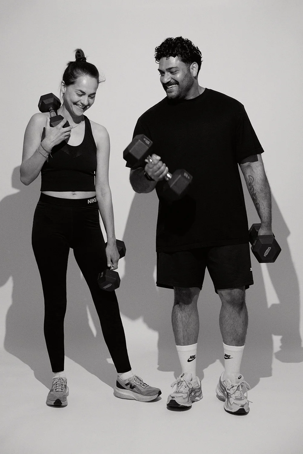A woman and a man holding dumbbells and smiling together in workout clothes, standing against a plain background.