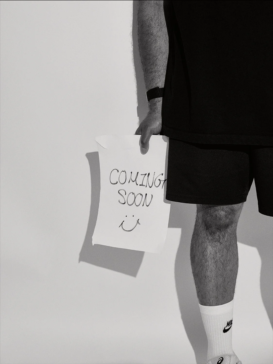 Person holding a sign that says 'COMING SOON' with a smiley face, wearing black shorts, white Nike socks, and a wristwatch.