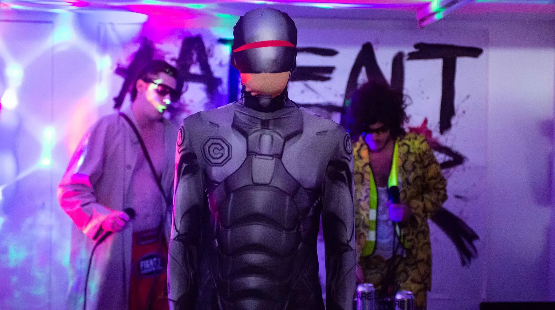 Person in a futuristic gray suit and helmet standing amidst other people at a performance with colorful lighting.