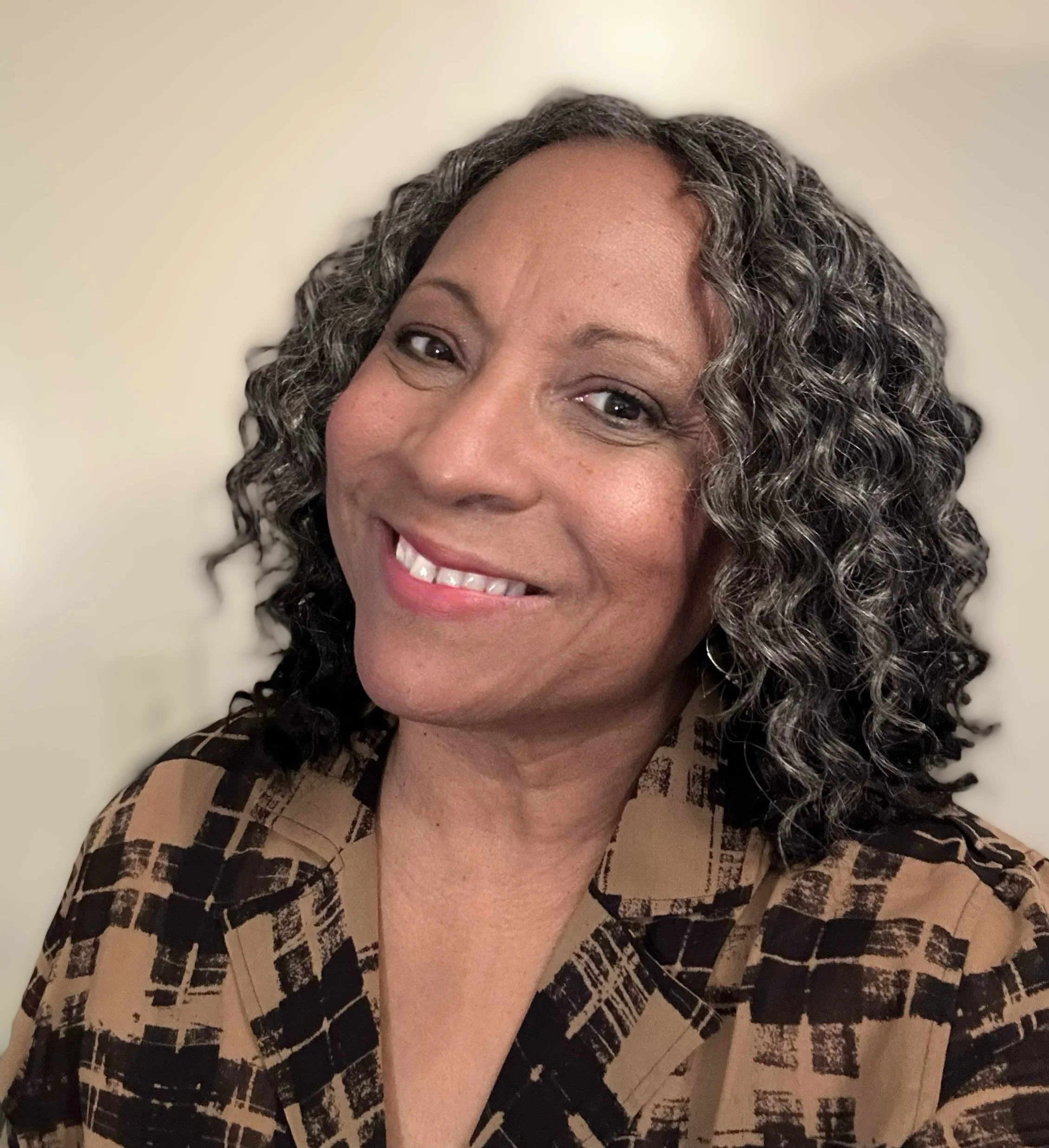Portrait of a woman with curly hair smiling at the camera, wearing a patterned shirt against a plain background.