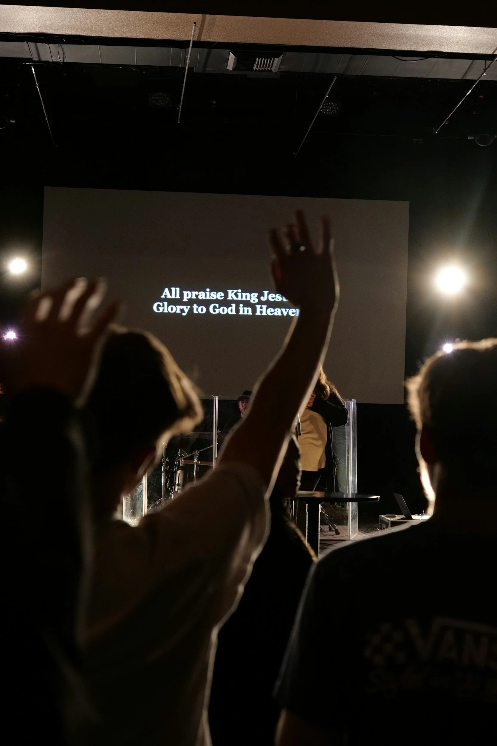 People raising their hands in a church service or religious gathering, with a large screen displaying lyrics in the background.