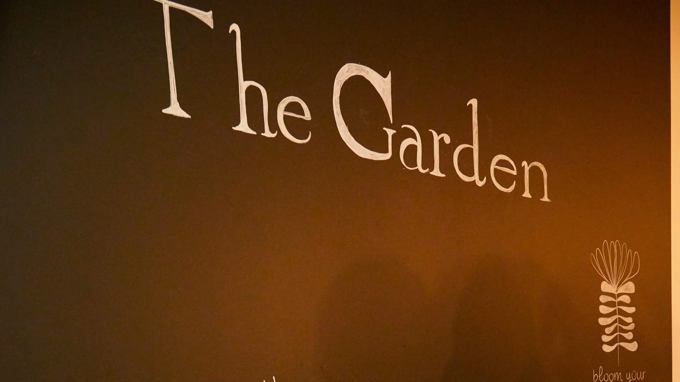 Scritta 'the Garden' su una lavagna con un disegno stilizzato di un fiore o pianta in basso a destra e la scritta 'bloom your balance'.
