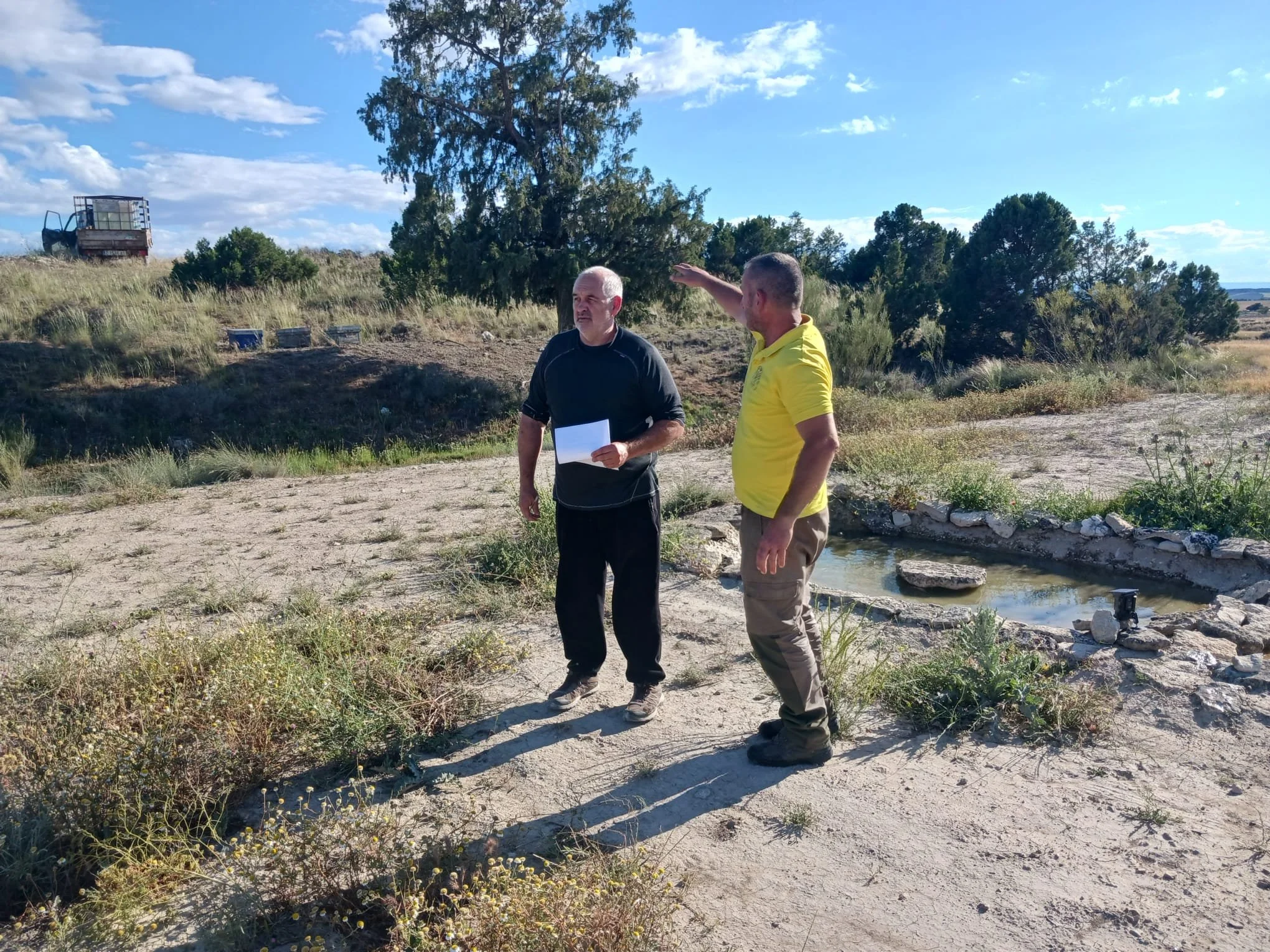 Two men are standing outdoors on a sunny day, having a conversation near a small pond or water feature surrounded by rocks. One man, dressed in black, is holding papers and appears to be listening, while the other man, in yellow, is pointing towards something in the distance. Trees and a clear sky are visible in the background.