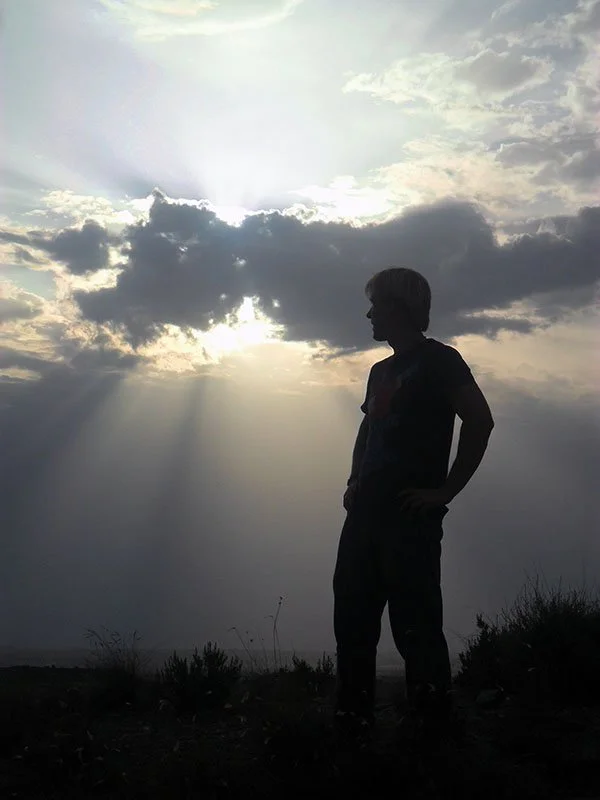 Silhouette of a boy standing outdoors during sunset or sunrise, with clouds in the sky and rays of sunlight shining through.