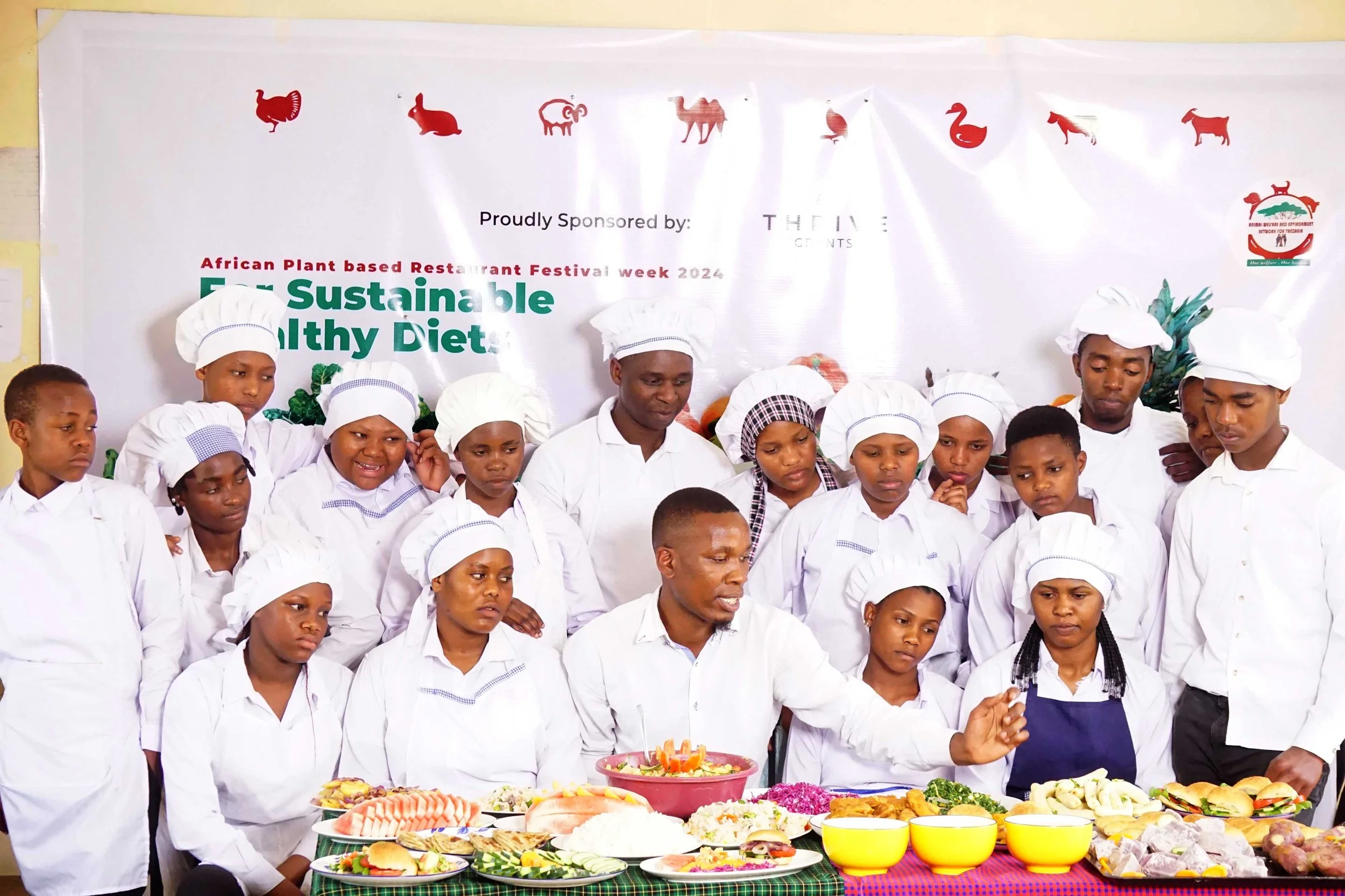 Group of children and adult chefs in white chef hats and coats, gathered around a table with various dishes and ingredients during a culinary event.