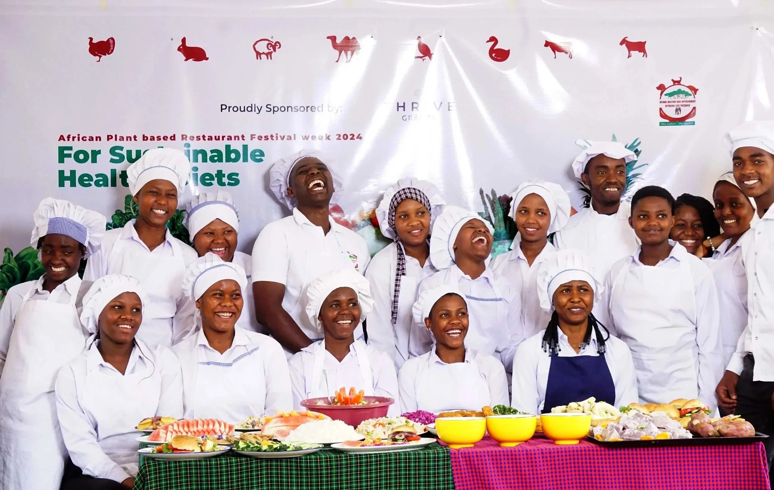A group of diverse chef adults and children in white chef uniforms, smiling and laughing, standing behind a table filled with various traditional African dishes and ingredients, at the African Plant Based Restaurant Festival.