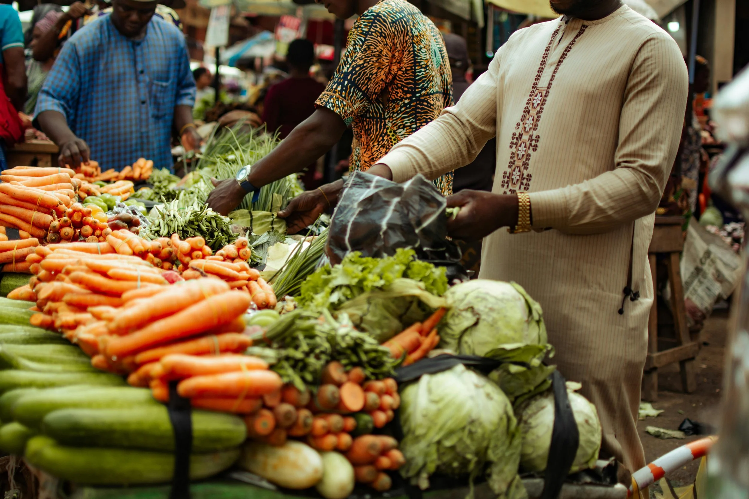 People shopping for fresh vegetables at an outdoor market, with carrots, cabbages, and other produce displayed on the stand.