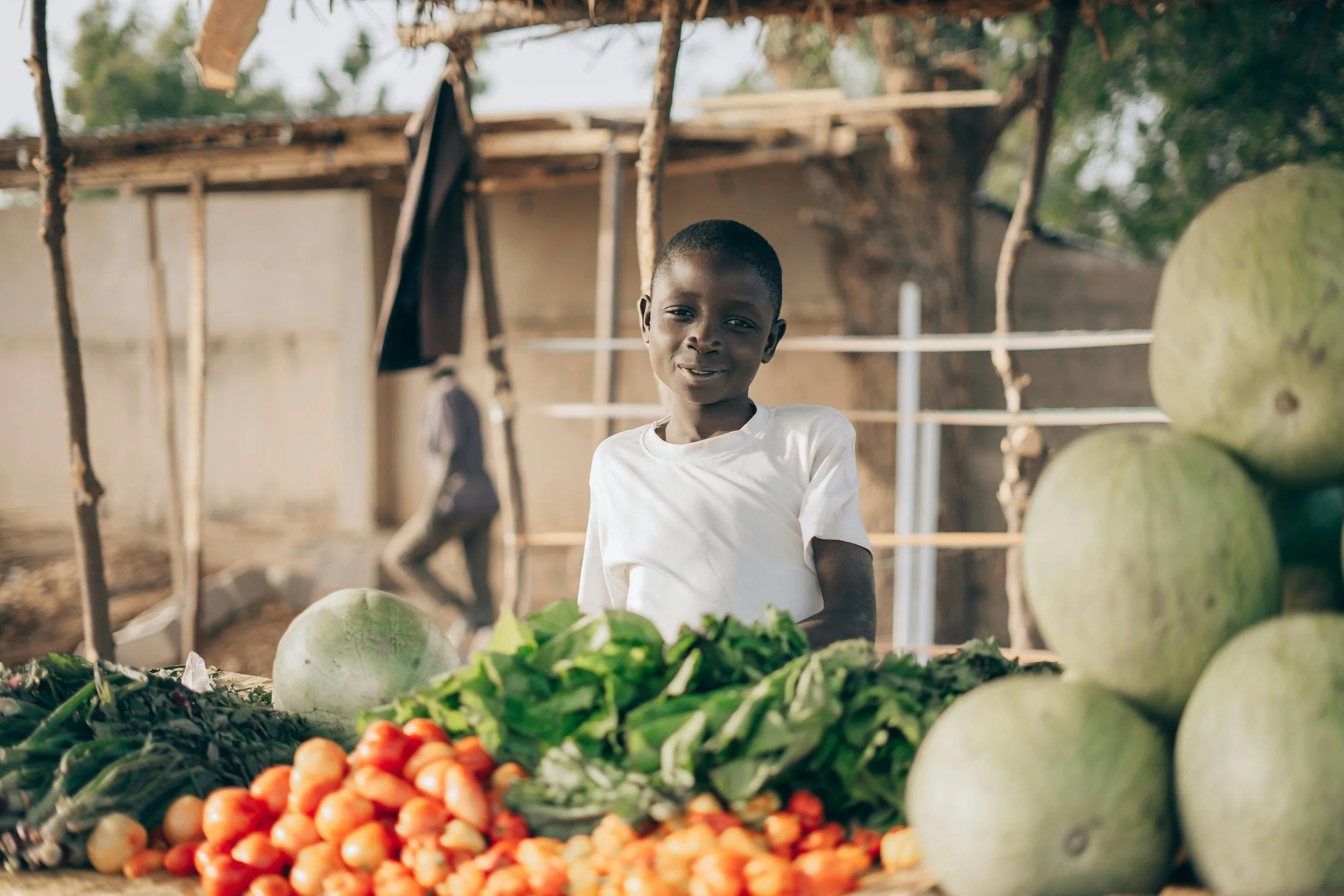 A young boy standing behind a display of fresh vegetables at an outdoor market, with large melons and leafy greens visible.