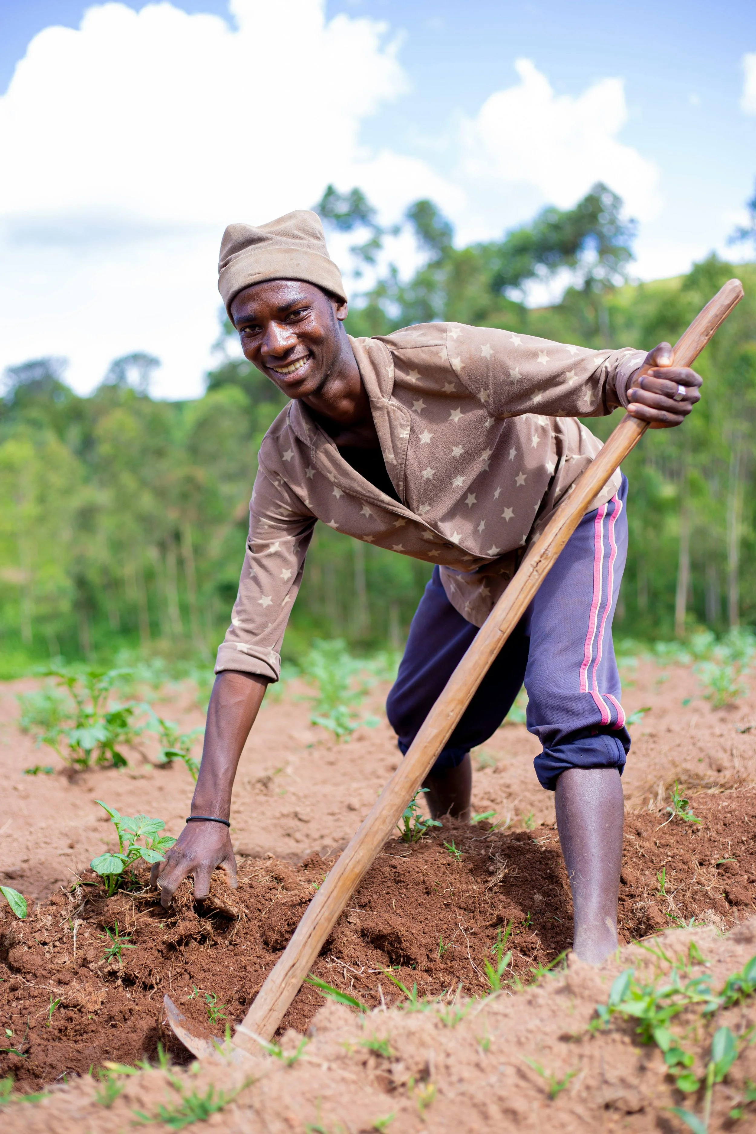 A young man farming, wearing a beige beanie, working with a hoe in a field of small green plants, with trees and a cloudy sky in the background.