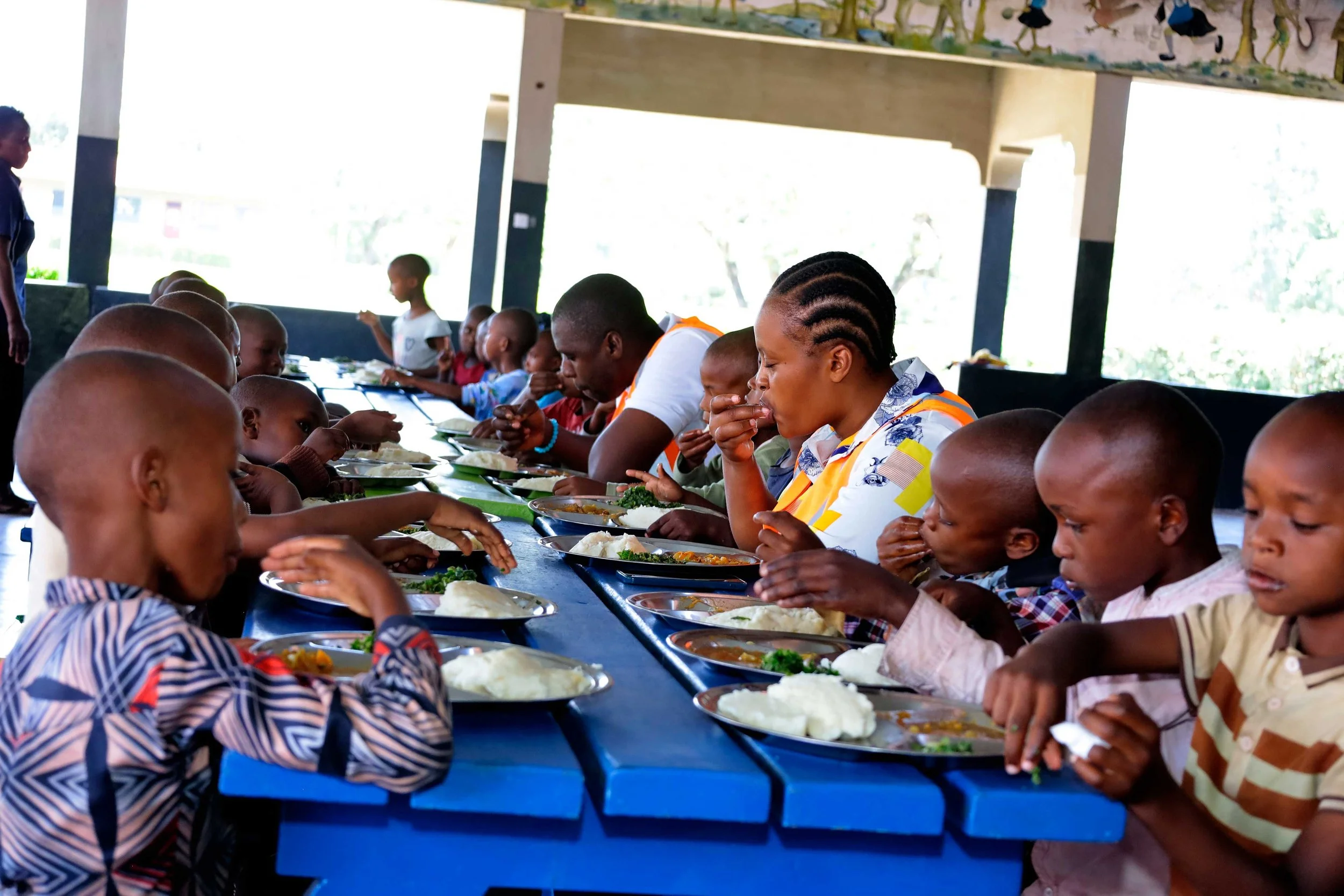 Children and adults sitting at a long blue table, eating a meal with plates of food including rice, vegetables, and soup, inside a sheltered building with windows and some artwork or decorations on the walls.