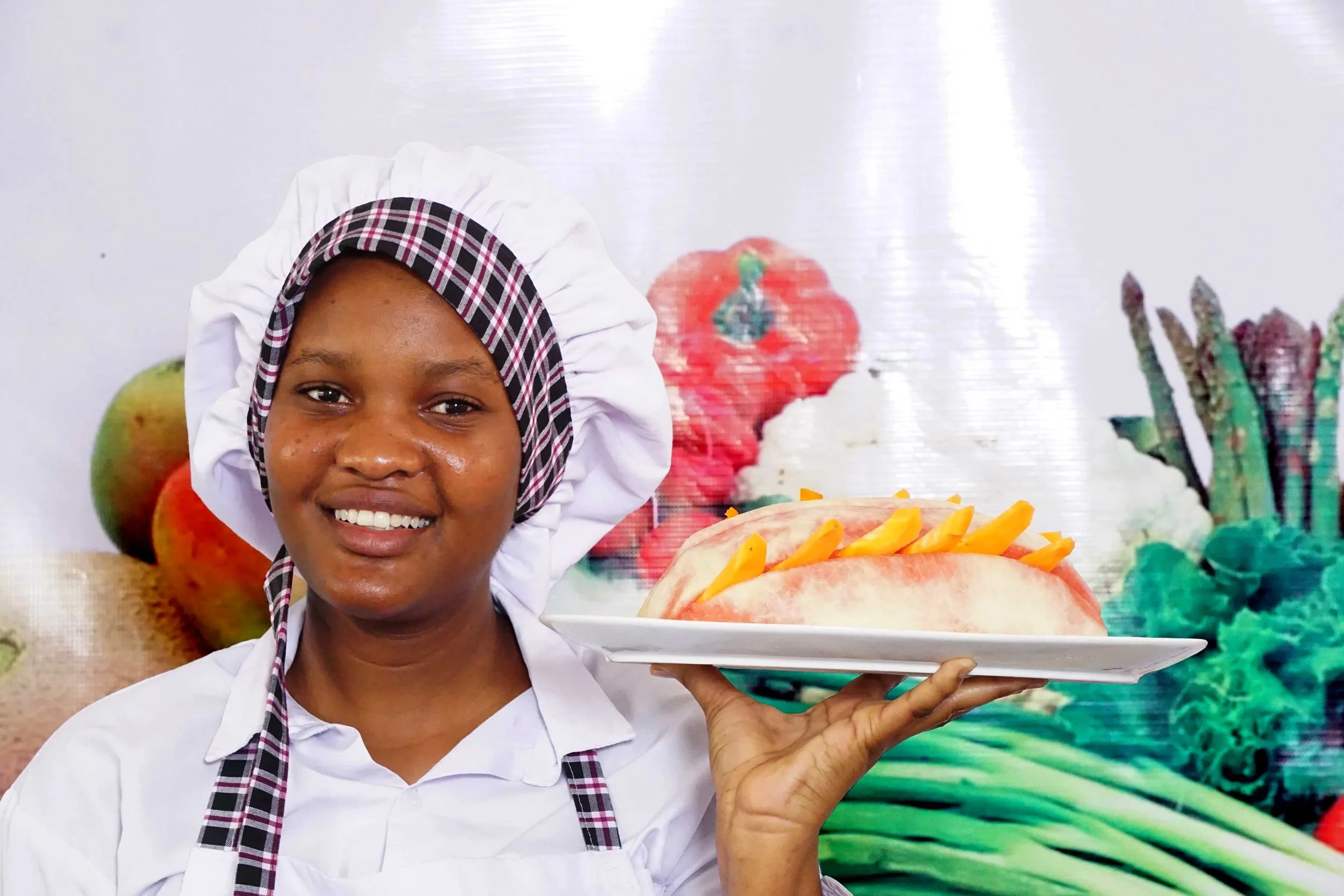 A smiling female chef in a white uniform and checkered headscarf holding a plate of sliced watermelon with mango on top, standing in front of a colorful fruit and vegetable background.