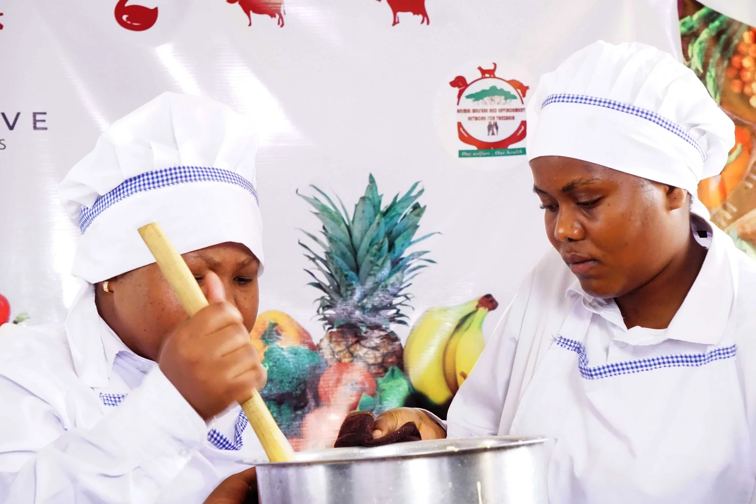 Two women wearing white chef uniforms and hats stirring a large pot, with a tropical fruit background including pineapple, bananas, and other fruits.