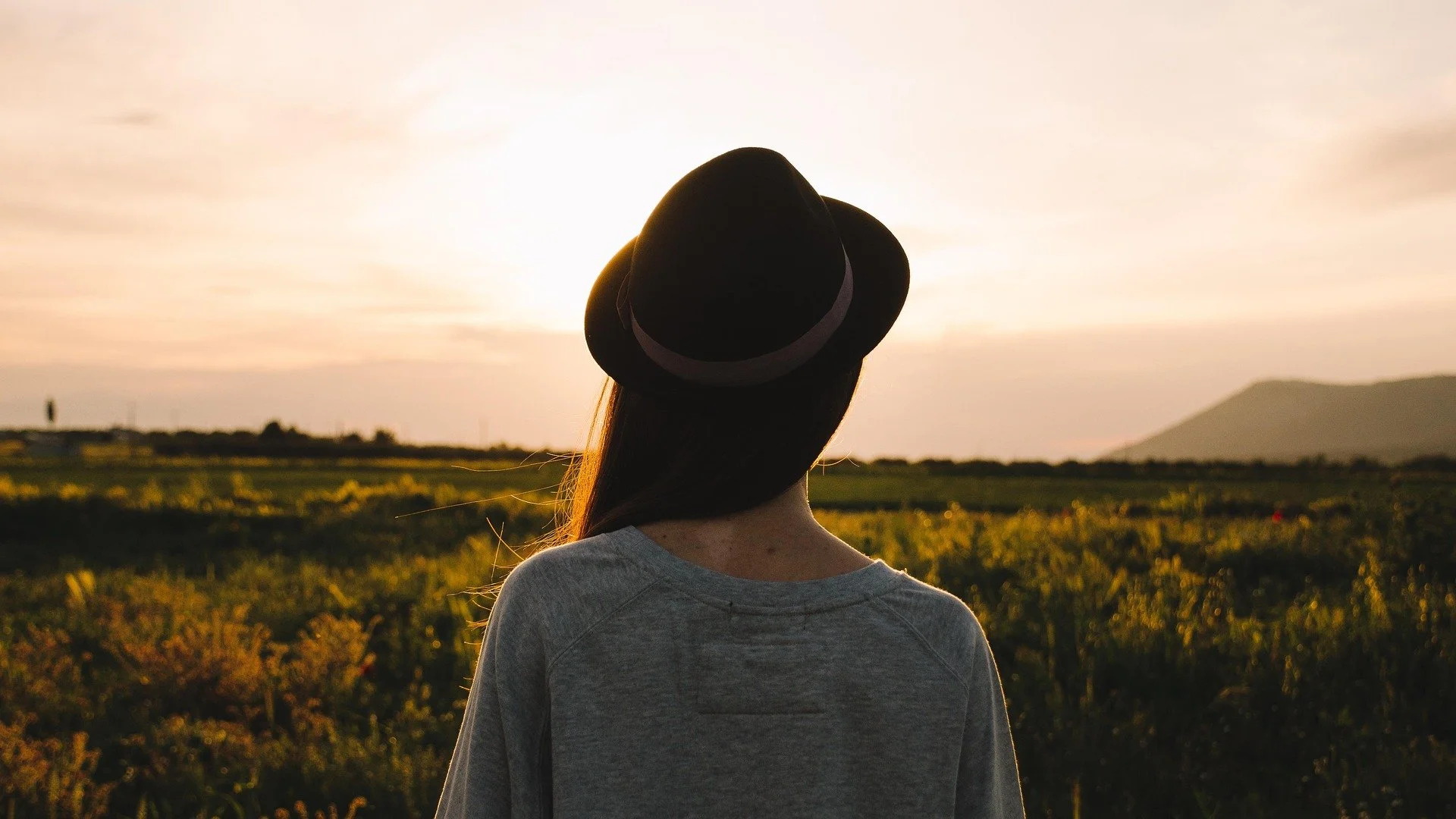 A woman with long hair wearing a black hat and gray sweatshirt looking at a sunset over a field with mountains in the distance.