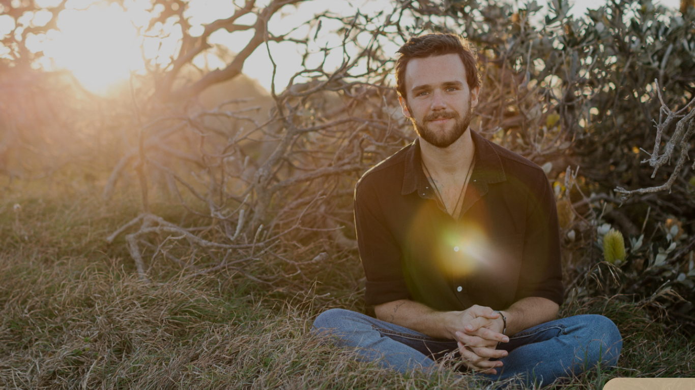A man with dark hair and a beard, sitting cross-legged on grass in front of a tree, during sunset with sunlight creating a flare.