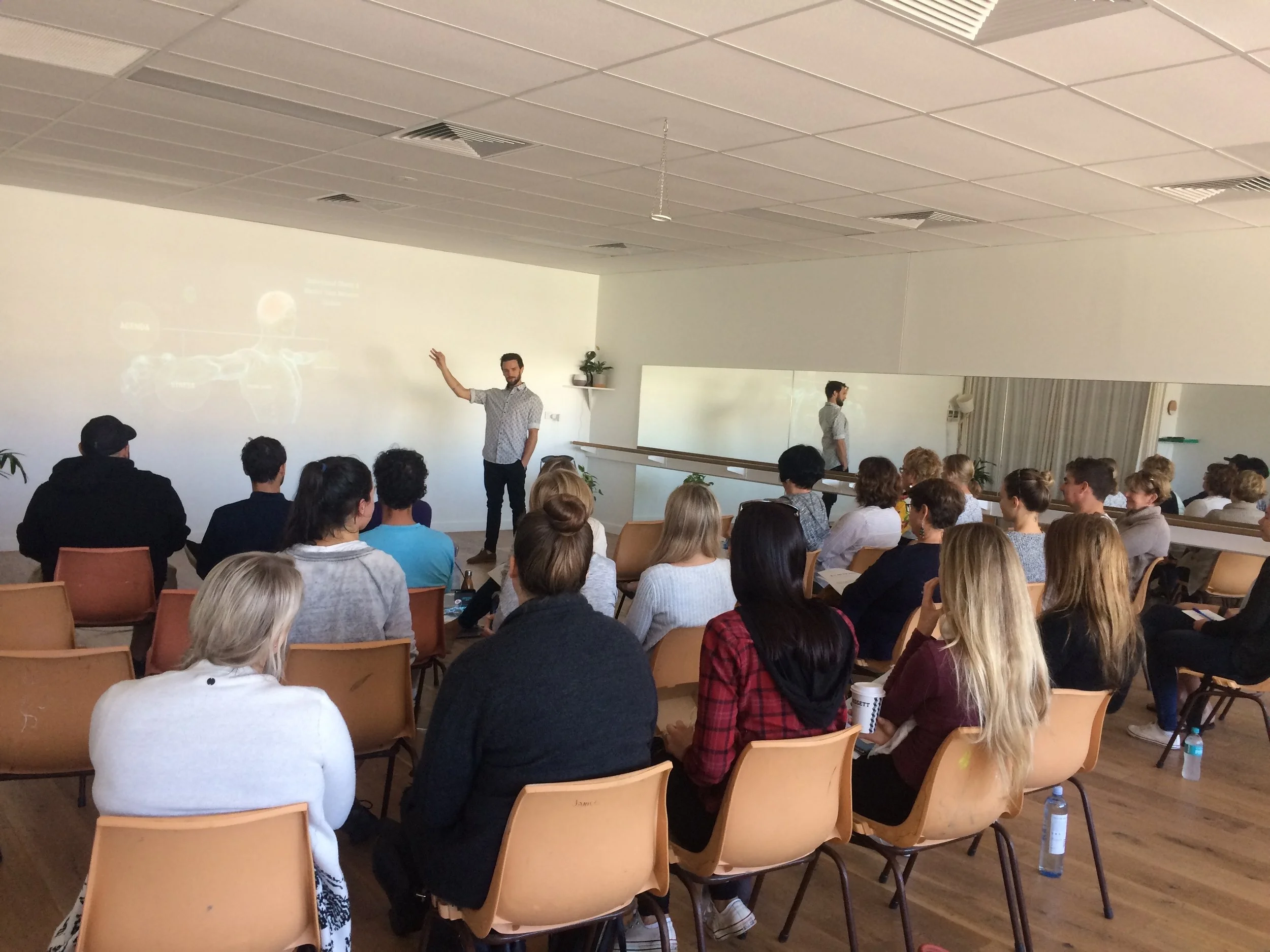 A man is giving a lecture or presentation to a seated audience in a classroom or conference room. The speaker stands in front of a white wall, pointing at a projection or display above, while the attendees sit on chairs, some taking notes or drinking beverages. A mirror on the side wall reflects the room and the speaker.