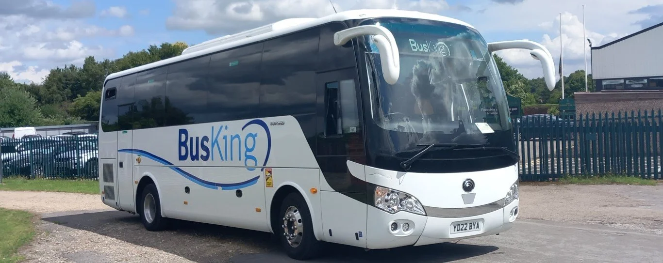 White BusKing coach bus parked outdoors on a gravel and asphalt surface, with a fence and trees in the background.