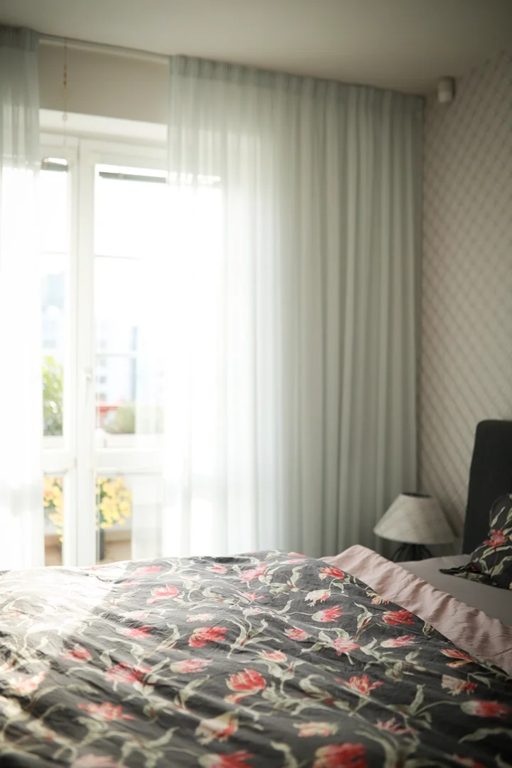 A bedroom with a bed that has a floral bedspread, a bedside lamp, and large windows with white curtains letting in sunlight.