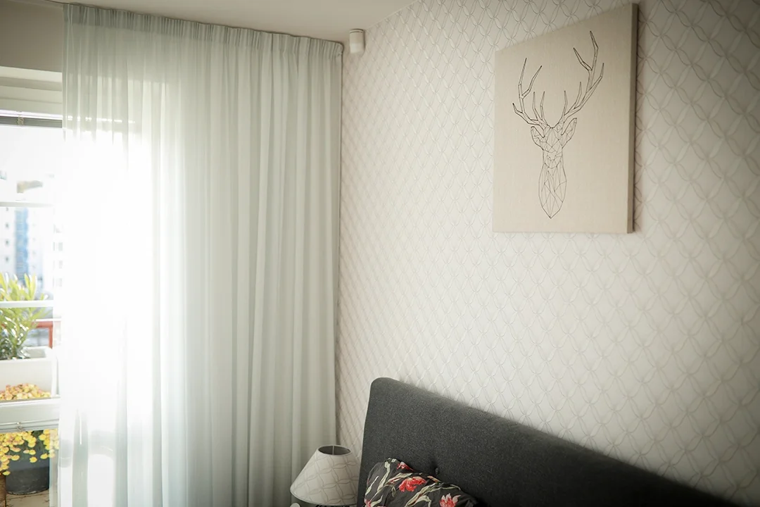A bedroom with white patterned wallpaper, a canvas of a geometric deer head on the wall, a deep gray headboard, and sheer white curtains covering a balcony door with plants outside.
