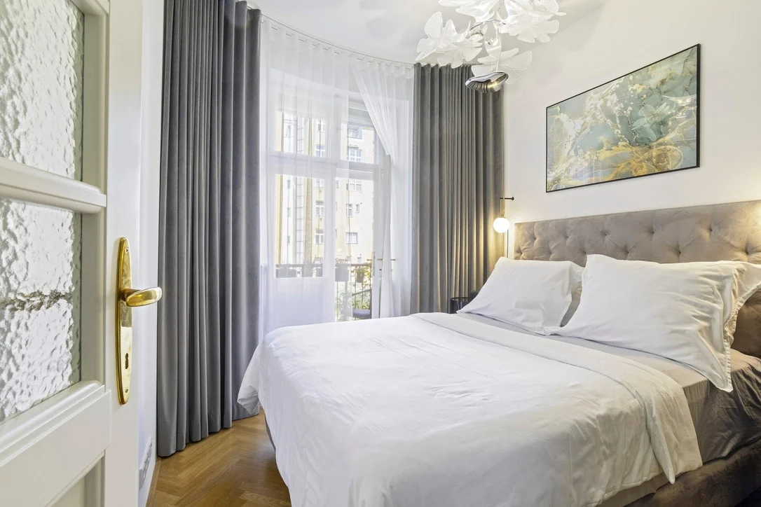 A bedroom with a large bed, gray tufted headboard, white bedding, and pillows, with a window covered by white sheer curtains and gray drapes, a nightstand with a lamp, a framed wall artwork, and a modern white chandelier.