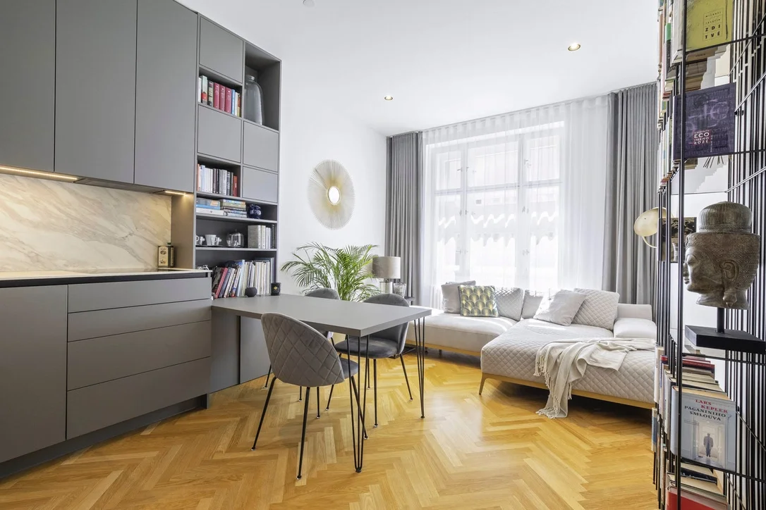 Bright living room with large window, white L-shaped sofa, gray dining chairs, gray cabinetry, open bookshelf, hardwood floor, decorative wall art, houseplants, and books.