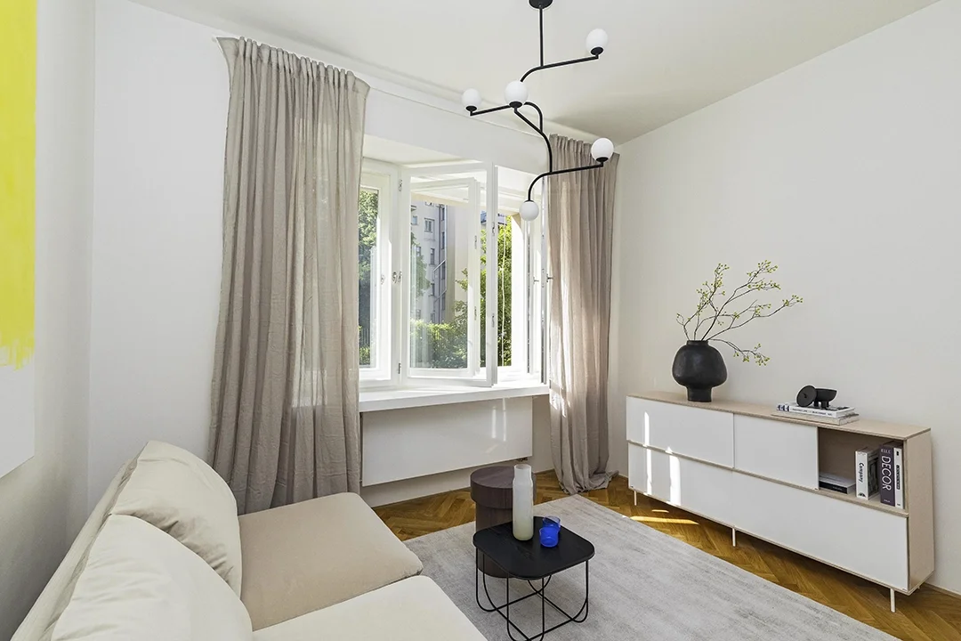 A living room with cream-colored sofa, a black side table with a white vase and a blue candle, large windows with beige curtains, a white sideboard with a black vase and books, and a modern black and white chandelier.
