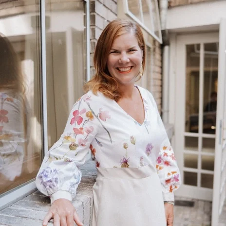 A smiling woman with shoulder-length red hair standing outside near a window and a door, wearing a white blouse with colorful floral embroidery.