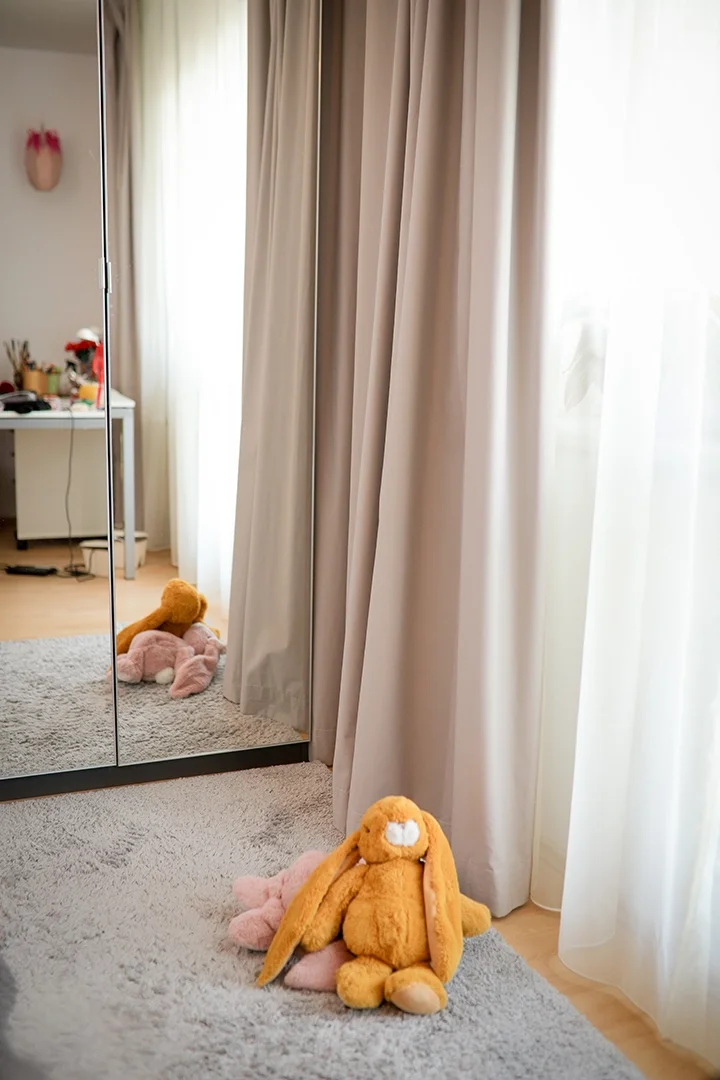 A plush orange bunny toy with long ears sitting on a light-colored rug, with a mirror reflecting part of the room and a curtain in the background.