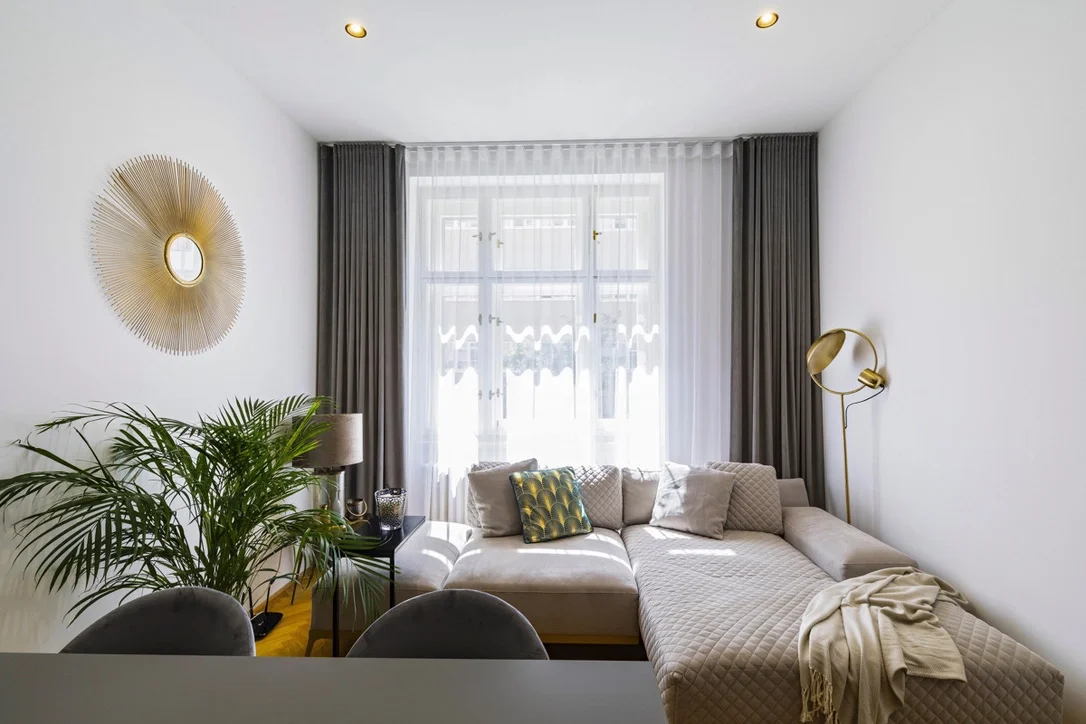 Bright living room with a beige sectional sofa, decorative pillows, and dark curtains framing a large window with sheer white curtains. A gold wall mirror and a gold floor lamp are on the walls, with a green potted plant and a side table with lamp and decorative objects nearby.