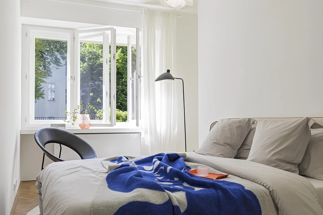 Bright bedroom with open window showing green trees, white curtain, bed with white sheets, beige pillows, a black chair, a floor lamp, and a blanket on the bed