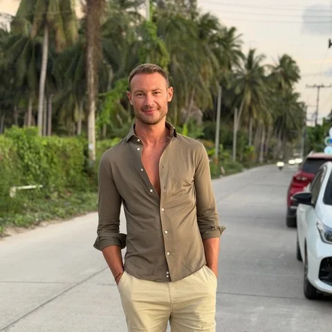 A man standing on a street lined with palm trees, with cars parked along the side, smiling and wearing a two-tone button-up shirt and light-colored pants.