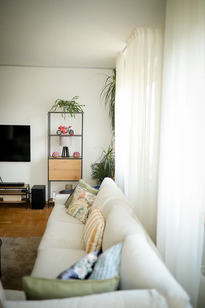 Living room with a white sofa, colorful pillows, a flat-screen TV, a black metal and wood shelf with a toy motorcycle, plants, and sheer white curtains.